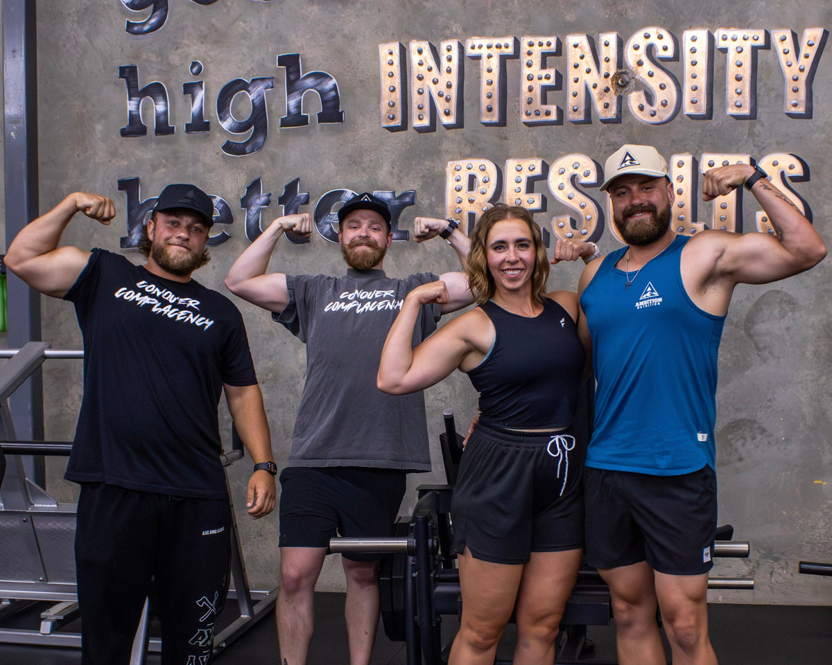 Four gym-goers posing with flexed arms in a gym setting, with motivational wall text in the background.
